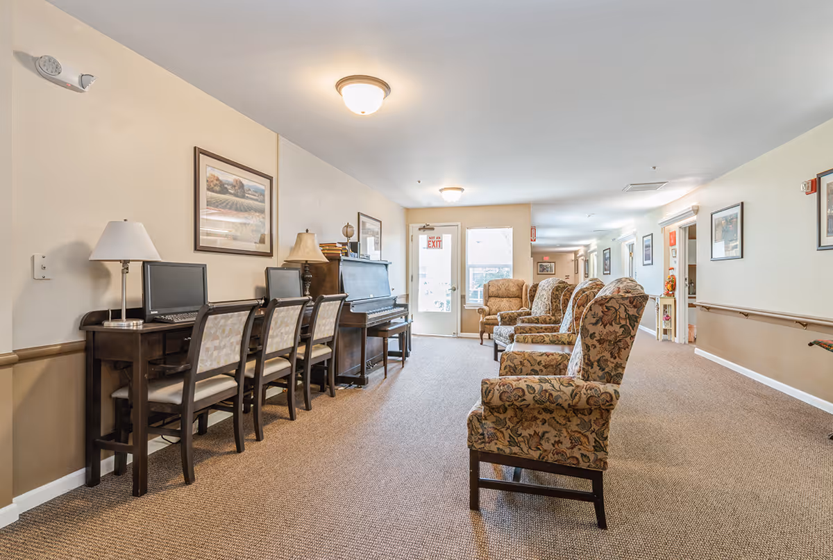 A well-lit common area in a senior living facility featuring a row of four computer desks with chairs and desktop computers on the left side, a piano with a lamp on top, and several upholstered armchairs with floral patterns arranged along the right side. The walls are decorated with framed artwork, and there is a door with a 'Not an Exit' sign at the far end of the room.