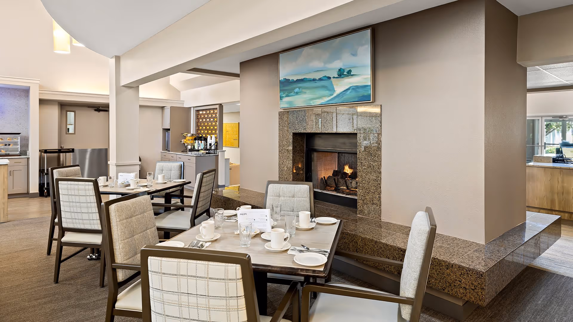A cozy dining area in a senior living facility with tables set for meals, featuring chairs with plaid upholstery. A lit fireplace with a granite surround is centrally located, and a landscape painting hangs above it. The room has neutral-colored walls and carpeting, with natural light coming from windows in the background.
