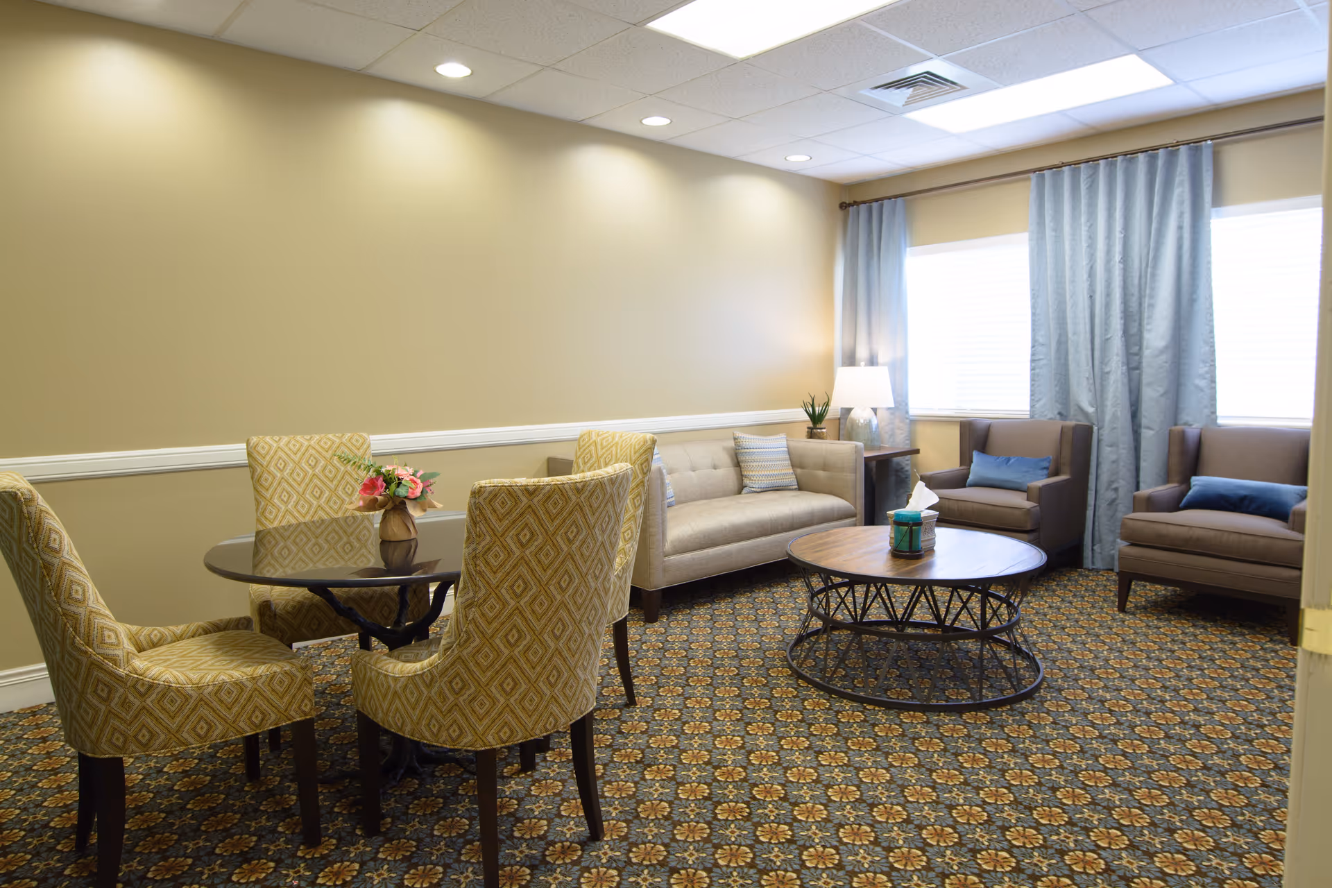 Well-lit sitting area with a sofa, armchairs, a round coffee table and a small dining table with four patterned chairs.