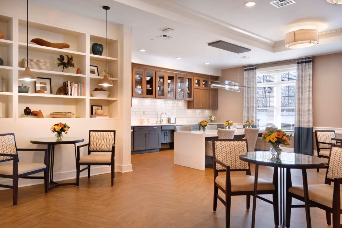 A bright and modern dining area in a senior living facility featuring round tables with chairs, each table decorated with a vase of colorful flowers. The room has a wooden floor, built-in shelves with decorative items and books, and a kitchen area with wooden cabinets, a white tiled backsplash, and a large island with seating. Large windows with curtains allow natural light to fill the space.