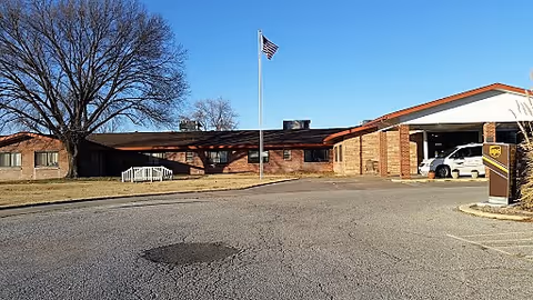Exterior view of a single-story brick building with a covered entrance where a UPS delivery van is parked. There is an American flag on a flagpole in front of the building, a large tree to the left, and a clear blue sky above.