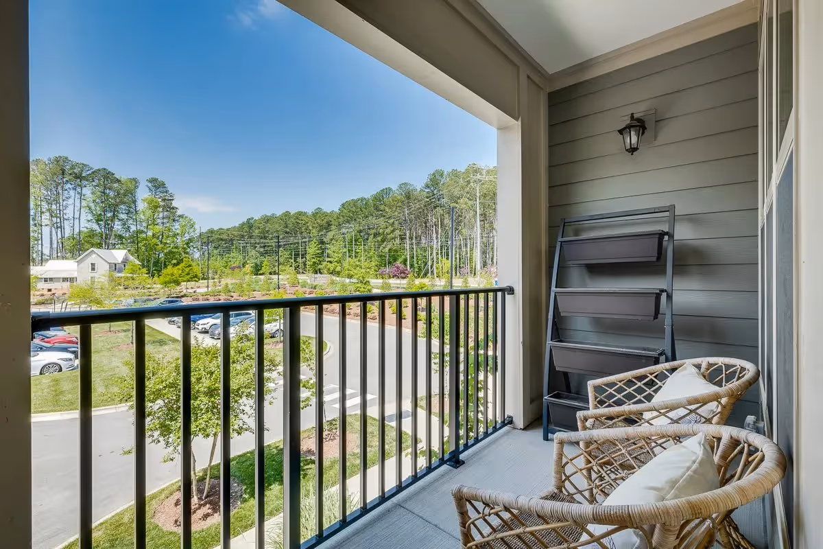Covered balcony with two wicker chairs and planters overlooking a parking lot and treeline.
