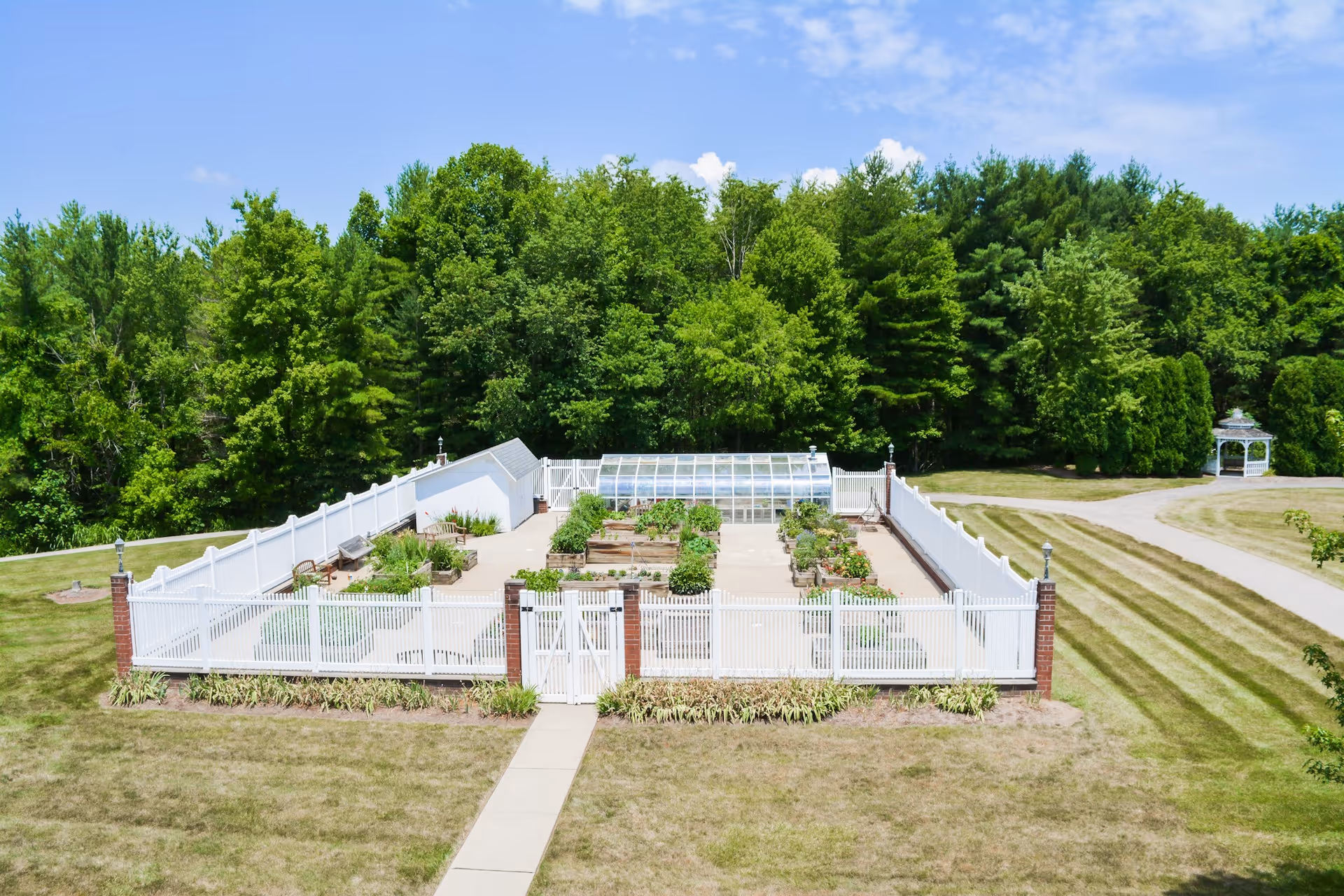 Walled outdoor community garden with raised beds and a small greenhouse, set on a lawn with trees in the background.