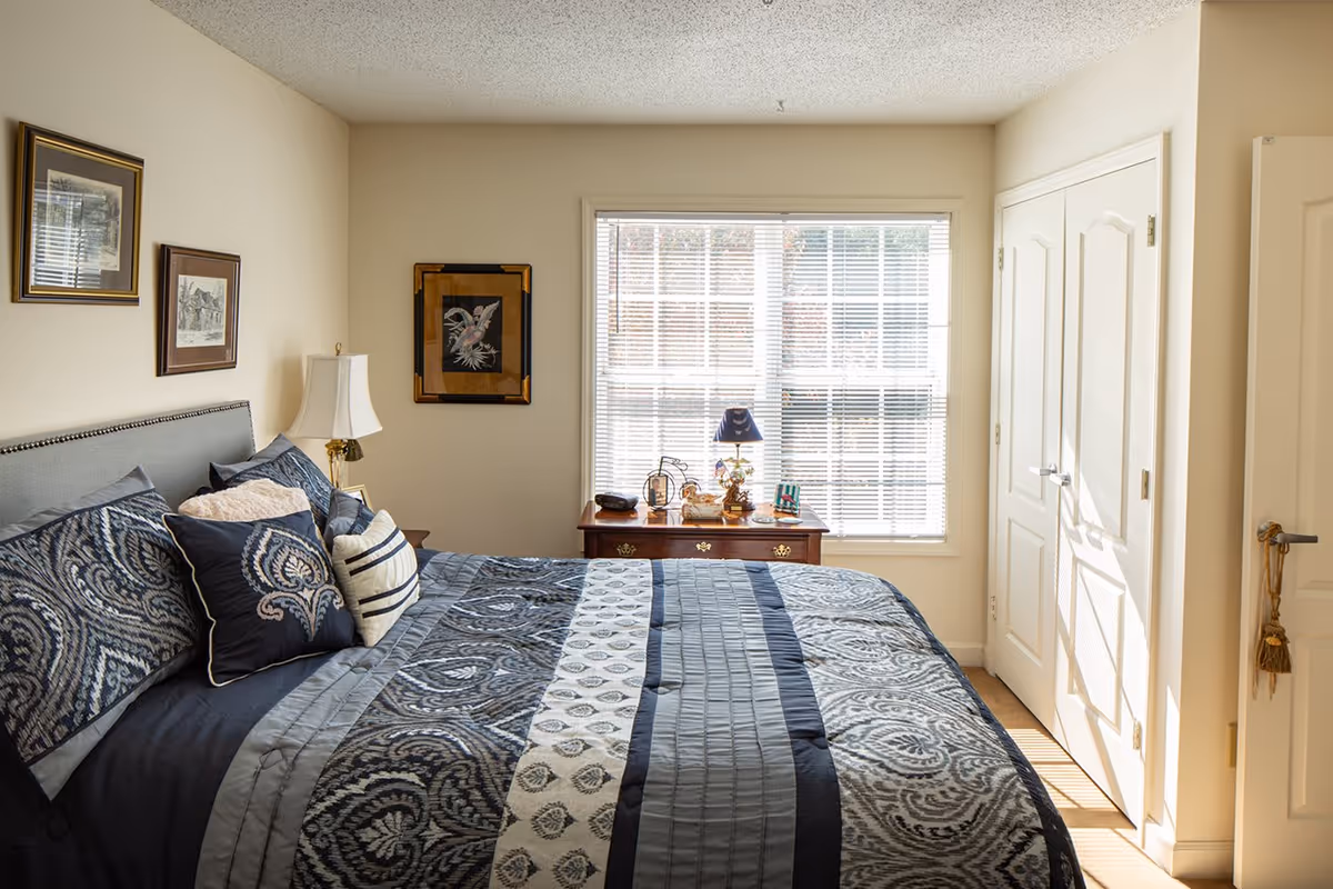 A cozy bedroom with a large bed covered in a patterned blue and gray comforter and multiple pillows. There is a wooden nightstand with a lamp and decorative items in front of a window with blinds. The walls are light-colored and decorated with framed artwork. A double-door closet is visible on the right side of the room.