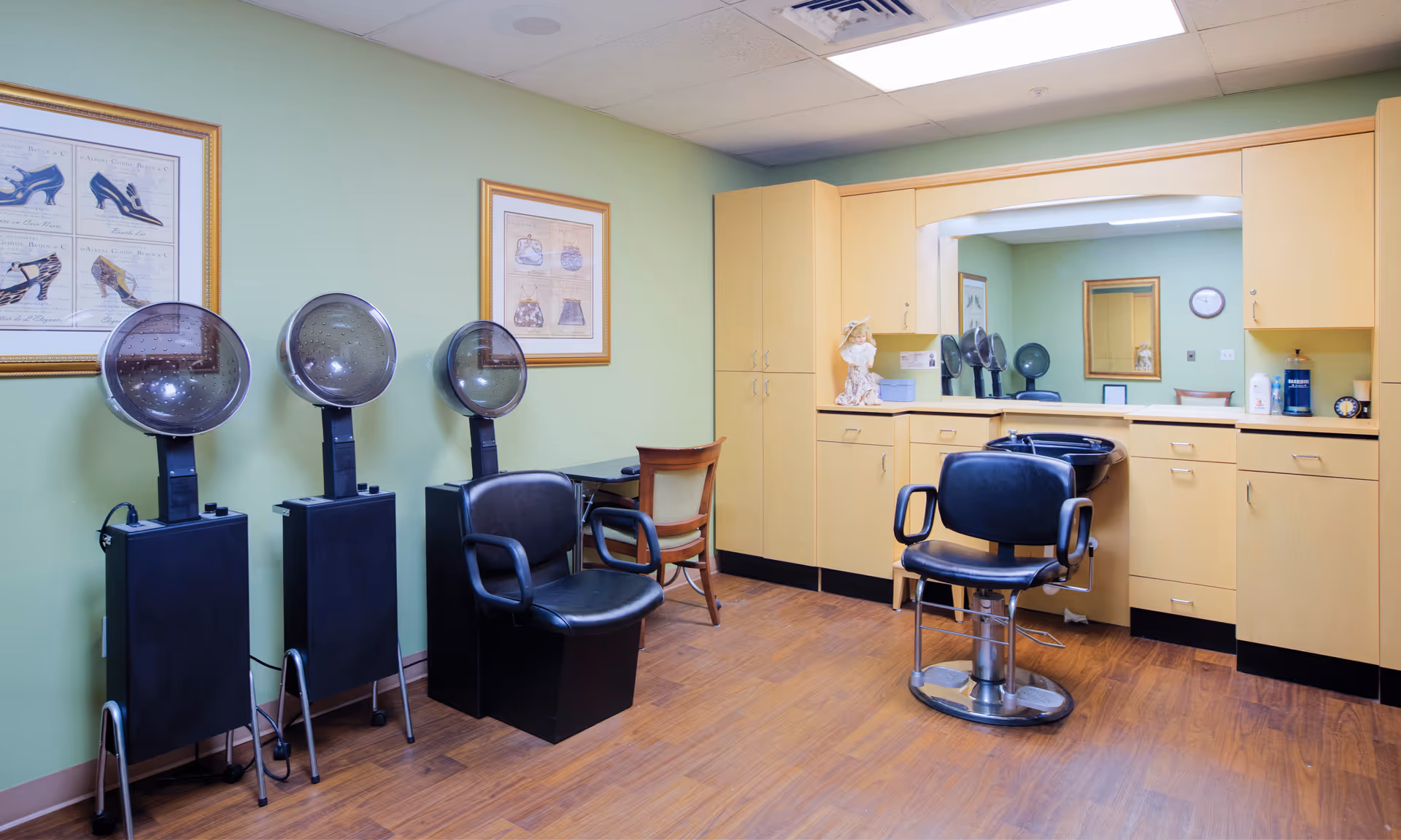 Interior of a salon area in a senior living facility with three black hair drying stations on the left, a black salon chair in front of a counter with cabinets and a large mirror, light green walls, wooden floor, and framed pictures on the wall.