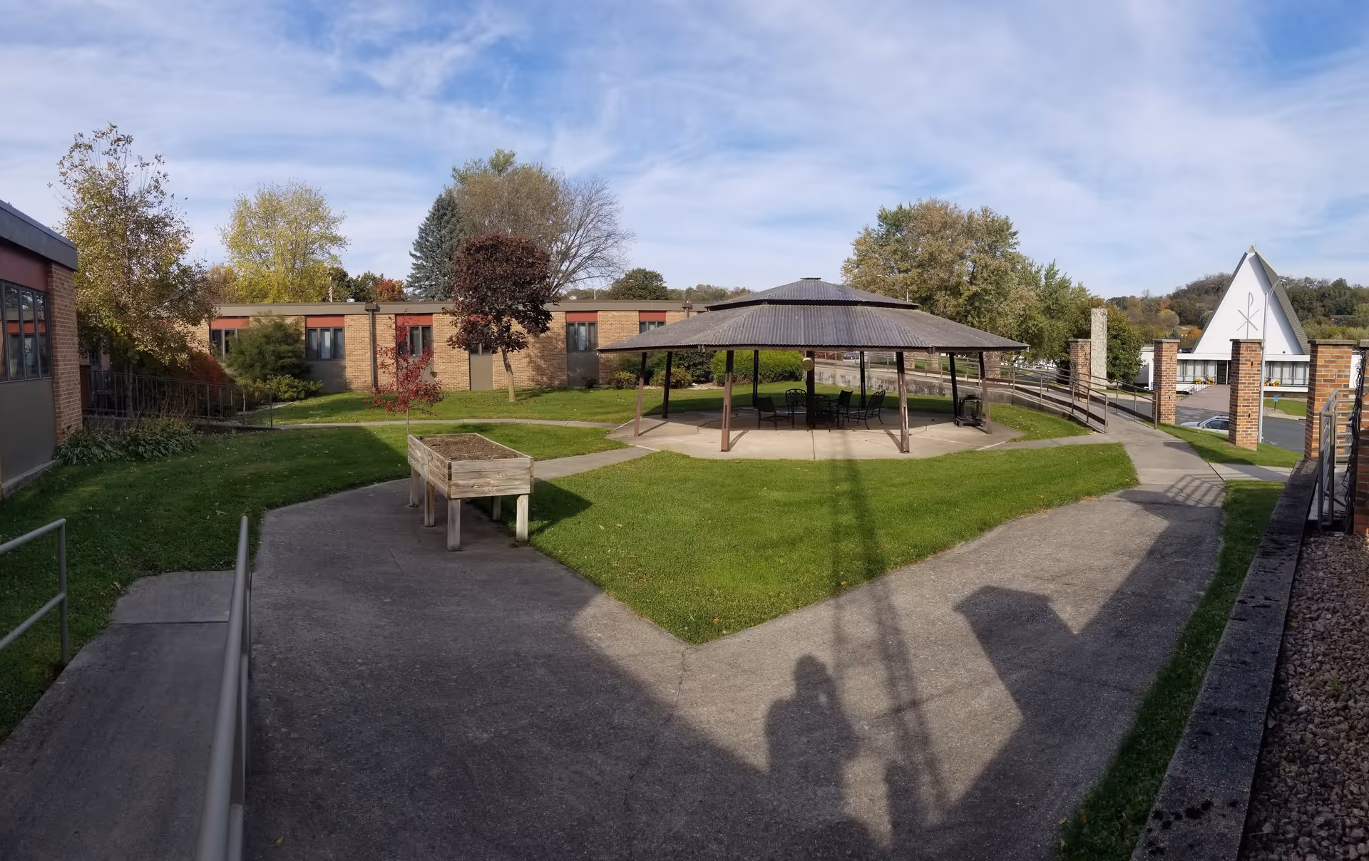 Outdoor courtyard area at Traditions of Preston featuring a covered pavilion with tables and chairs, surrounded by green grass and trees, with a brick building in the background and a church-like structure visible to the right.