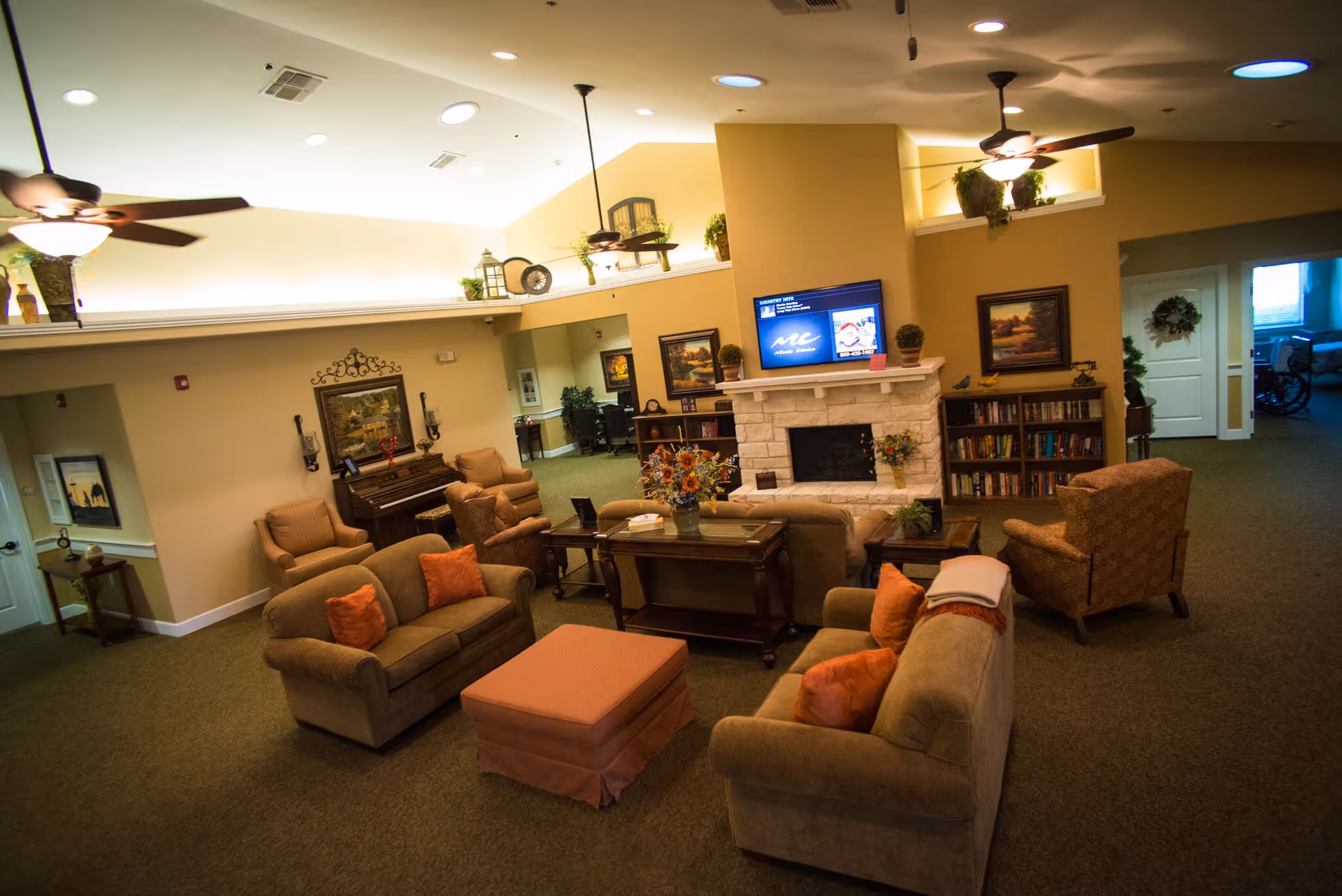 A cozy assisted living facility common area with multiple sofas and armchairs arranged around a central coffee table and ottoman. The room features a stone fireplace with a TV mounted above it, bookshelves filled with books, ceiling fans, and decorative plants and artwork on the walls. A piano is visible in the background along with a hallway leading to other rooms.