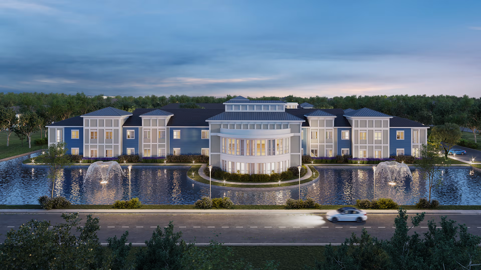 Front exterior view of The Meridian at Brandon facility at dusk, featuring a large building with multiple windows, a central rounded section, surrounded by a water feature with fountains, landscaped greenery, and a road with a car passing by in the foreground.