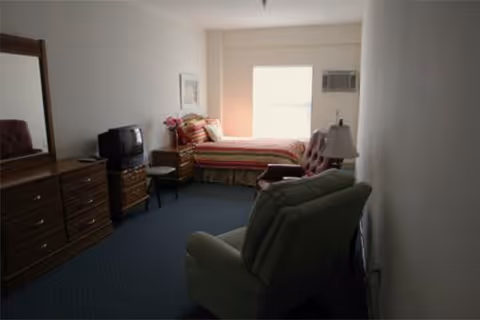 A simple senior suite with a single bed by a window, dresser with a TV, and two upholstered chairs.