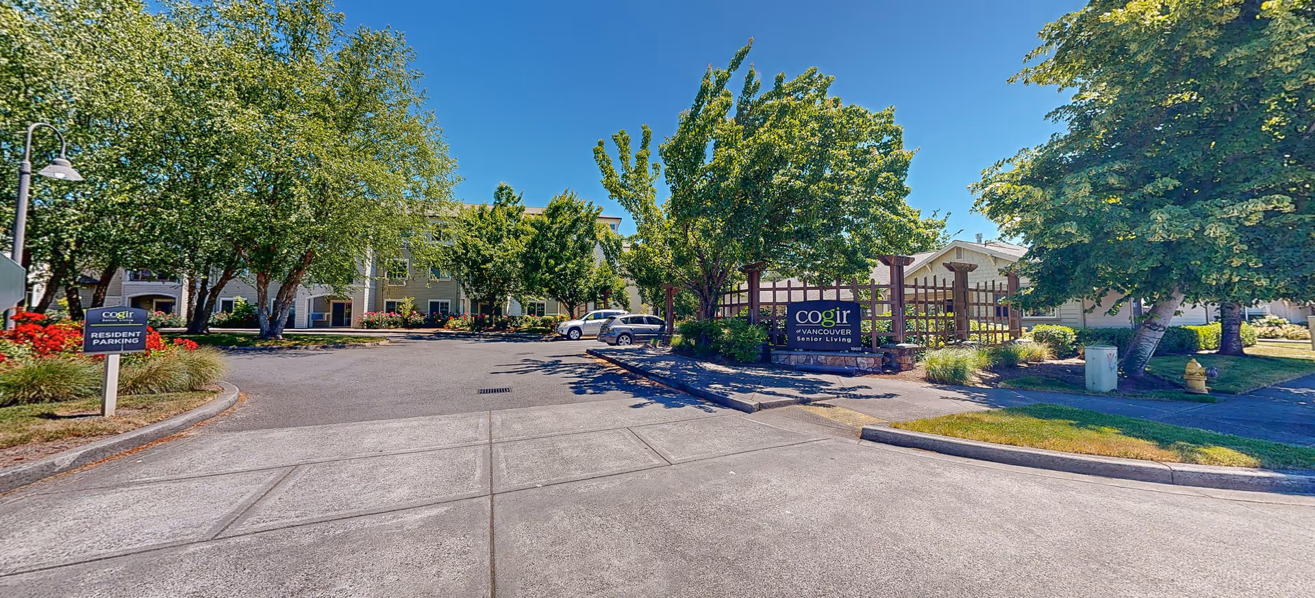 Exterior view of Brookdale Vancouver Orchards senior living facility showing a driveway entrance with resident parking sign, trees, and a building in the background under a clear blue sky.
