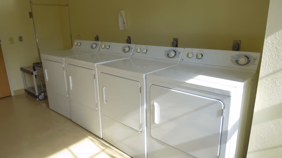 A laundry room with four white washing machines or dryers lined up against a pale yellow wall. There is a metal laundry cart with a white basket on the left side near a closed wooden door. Sunlight is streaming in from the right side, casting shadows on the machines and floor.