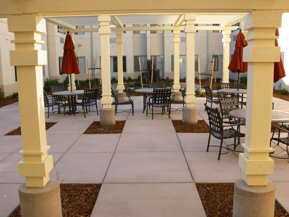 Outdoor patio area with multiple round metal tables and chairs arranged under a pergola with yellow columns. Red umbrellas are closed and positioned next to some tables. The patio is surrounded by a building with windows and some landscaping with plants and mulch.