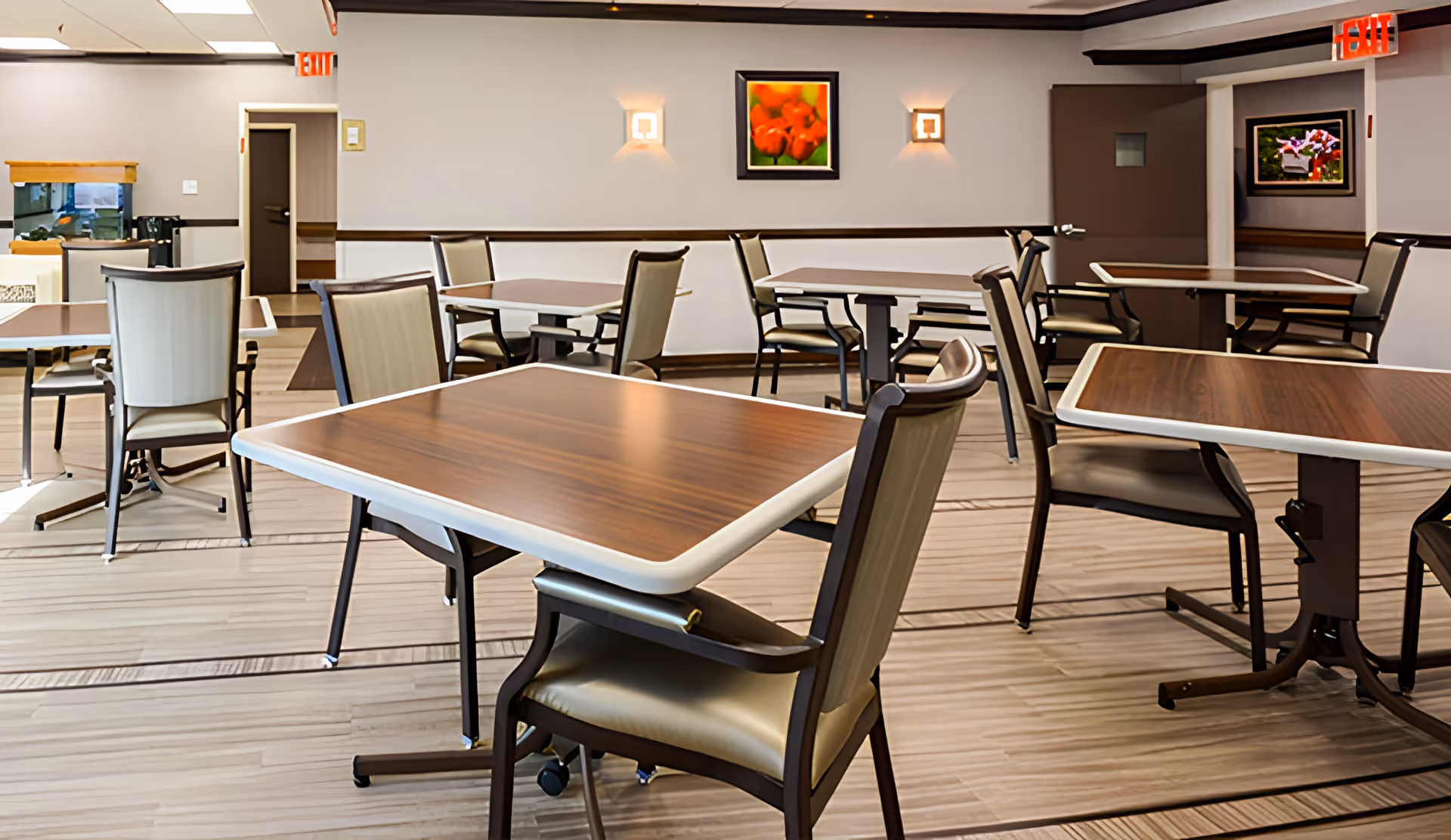 A clean and well-lit dining room with several square wooden tables and cushioned chairs arranged neatly. The room has light-colored walls adorned with framed floral artwork and wall sconces. There are exit signs above two doors in the background.