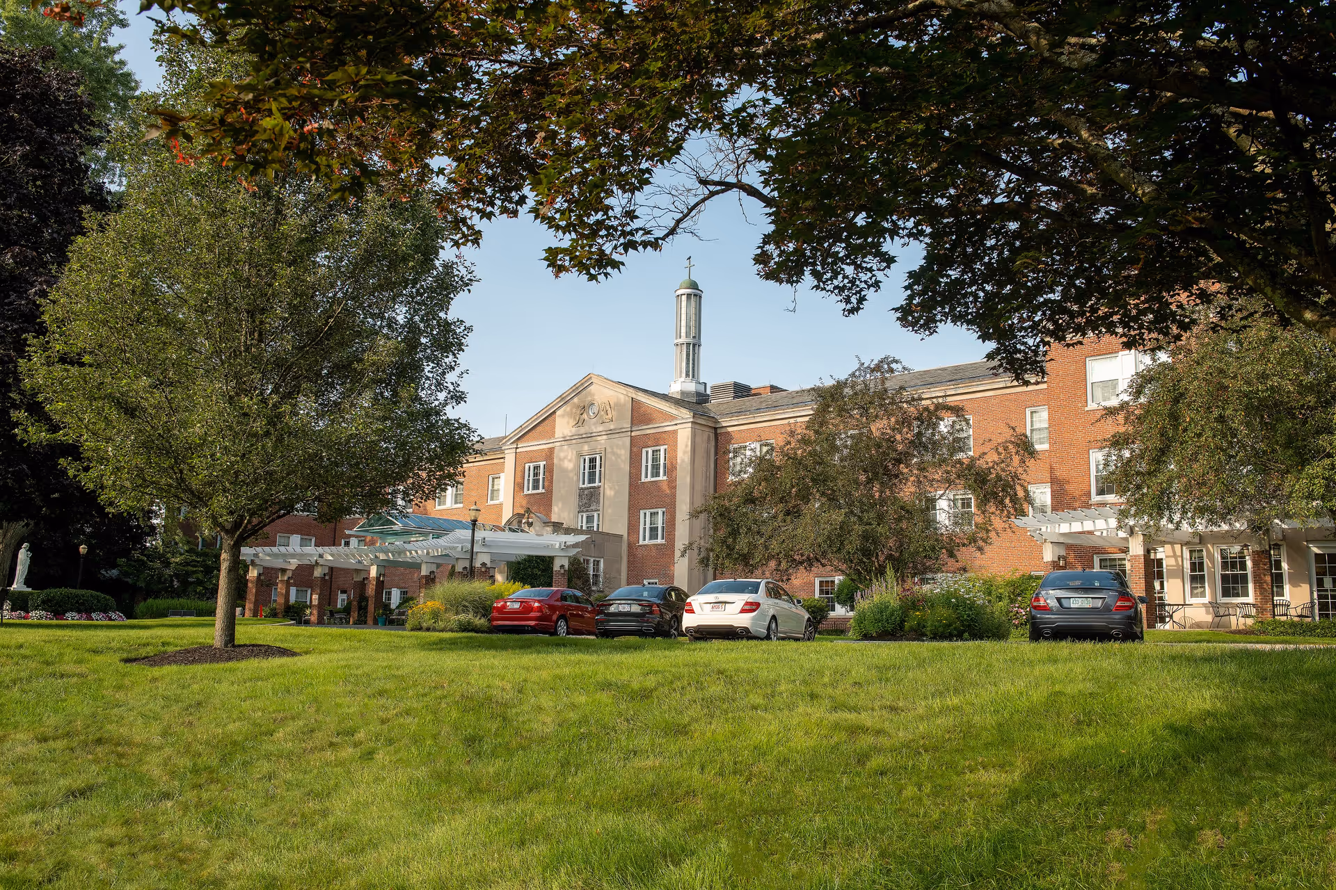 Exterior view of a large brick building with multiple windows, a central tower with a cross on top, and a white pergola entrance. Several cars are parked in front, surrounded by green grass, trees, and landscaping under a clear sky.