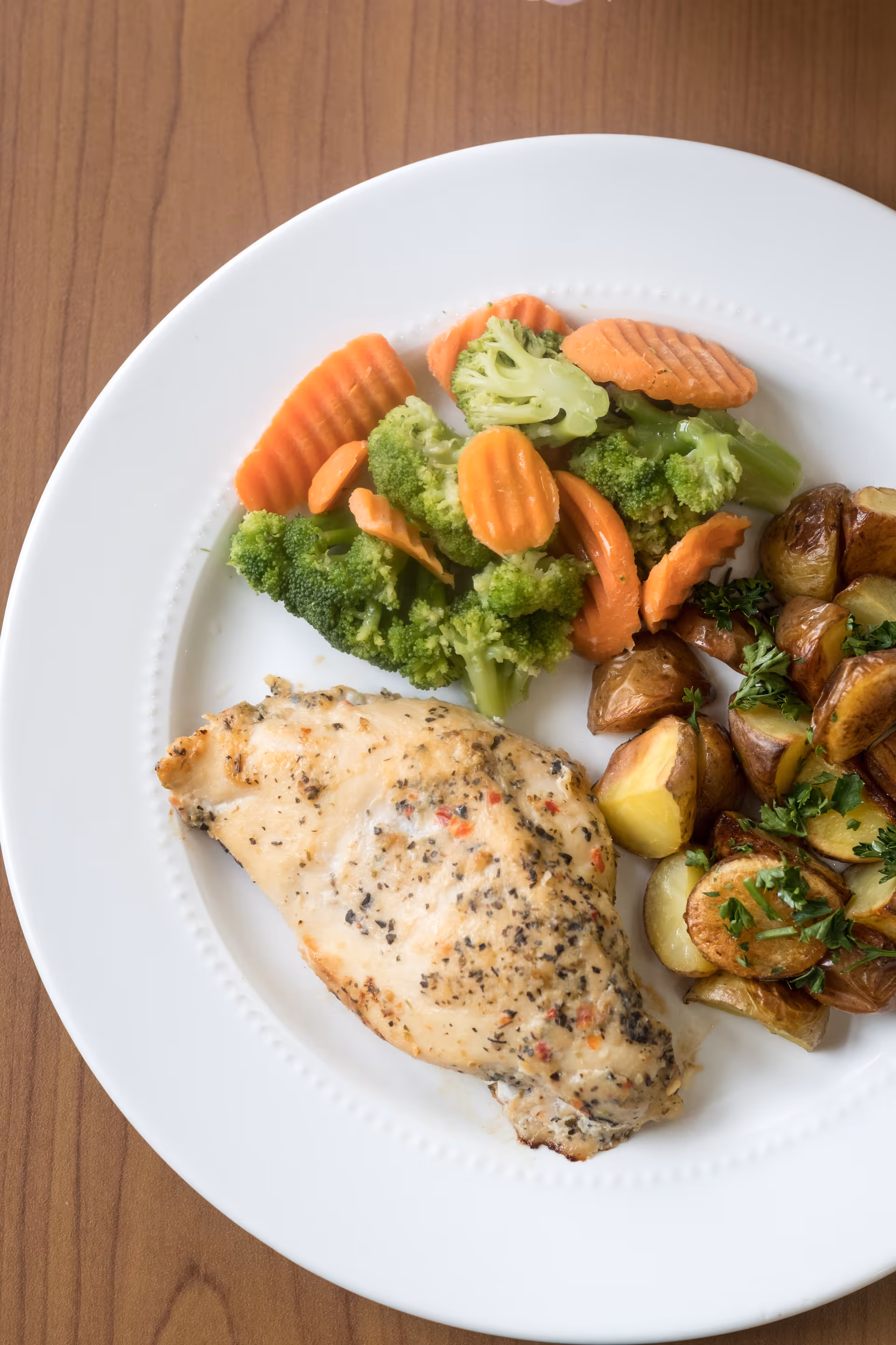 A white plate with a serving of seasoned grilled chicken breast, steamed broccoli and carrot slices, and roasted potatoes garnished with chopped parsley on a wooden table.