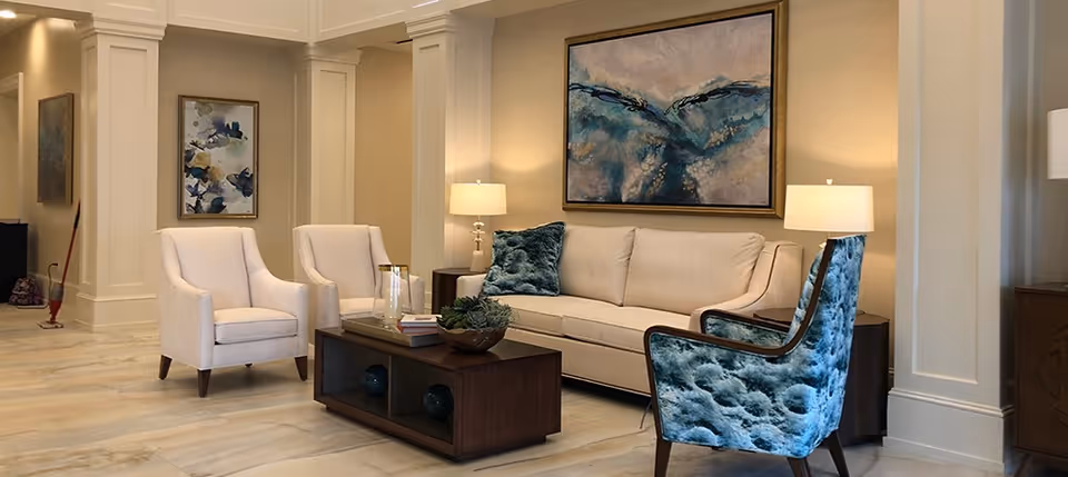 A cozy living room area with a beige sofa adorned with blue cushions, two white armchairs, and a blue patterned armchair. A wooden coffee table with decorative items sits in the center. The walls are beige with white trim, featuring two abstract paintings and two table lamps providing warm lighting.