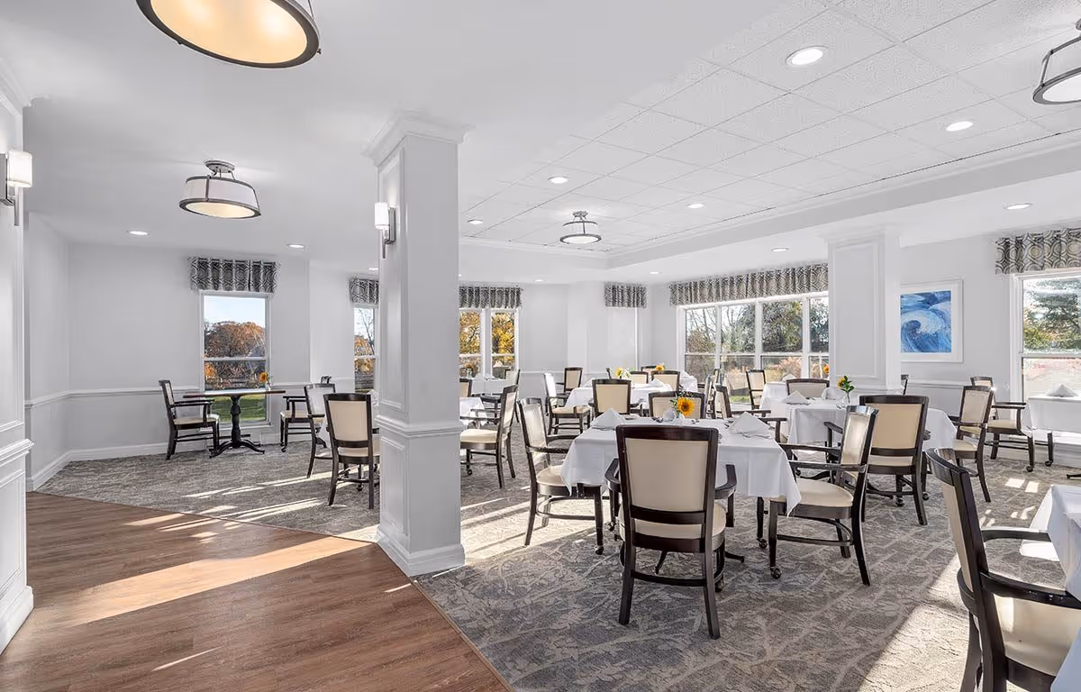 Bright airy dining room with multiple tables set with white tablecloths and chairs and large windows letting in natural light.