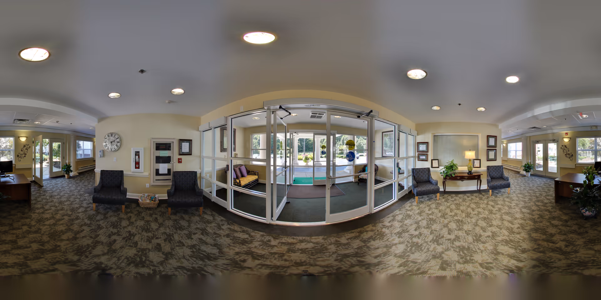 A spacious and well-lit senior living facility lobby with patterned carpet flooring, multiple seating areas with chairs, a small table with a lamp and framed pictures on the wall, large glass entrance doors leading outside, and windows allowing natural light to fill the room.