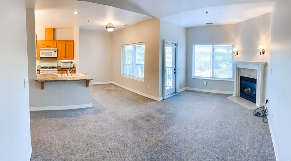 An empty open-concept living space featuring a kitchen with wooden cabinets, a granite countertop with a sink, and white appliances including a microwave and stove. Adjacent to the kitchen is a carpeted living room area with a fireplace, two wall-mounted light fixtures, a large window, and a glass door leading outside. The walls are painted light beige and the ceiling has recessed lighting and a hanging light fixture.