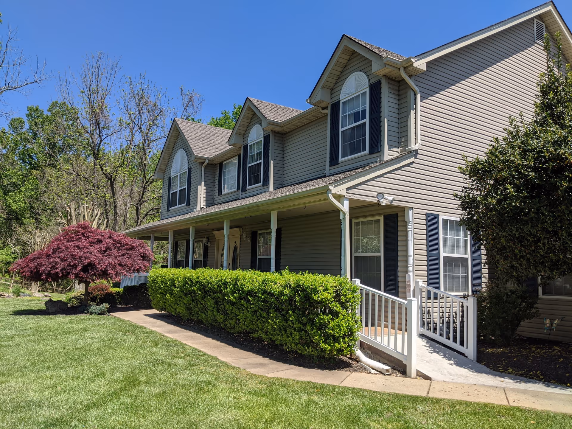 Two-story beige siding building with a covered front porch, accessibility ramp, manicured lawn, shrubs and a red ornamental tree under a clear blue sky.