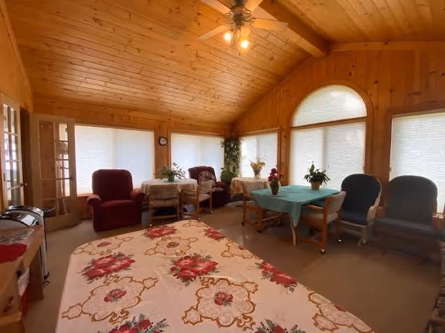 A cozy common area in a senior living facility with wooden walls and ceiling. The room features multiple tables covered with floral and solid tablecloths, surrounded by various chairs including armchairs and dining chairs. Large windows with blinds allow natural light to fill the space. There are plants and decorative items on the tables and in the corners of the room.