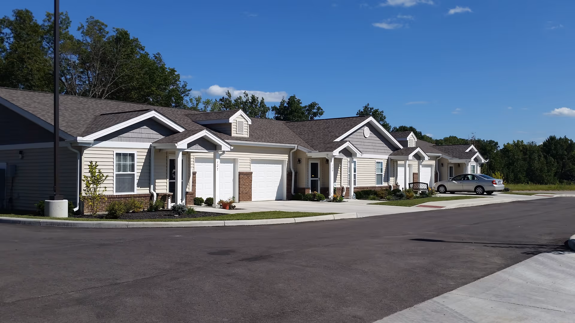 Exterior view of a row of single-story residential units with garages, driveways, and a parked car under a clear blue sky with trees in the background.