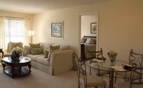A bright living and dining area with a beige sofa, round glass-top coffee table, floor lamp, and a small glass dining table with chairs, with a bedroom visible through an open doorway.