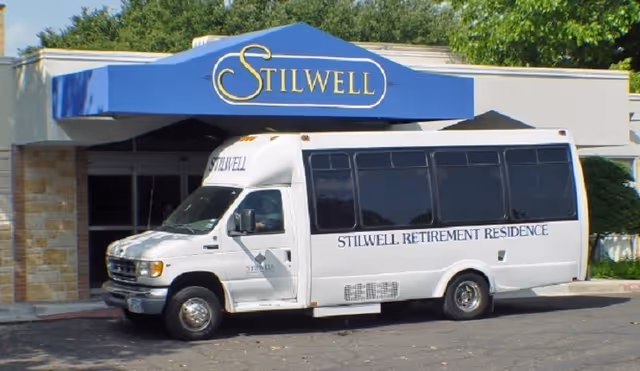 A white shuttle bus parked in front of the entrance to Stilwell Retirement Residence. The entrance has a blue awning with the word 'STILWELL' in gold letters. The bus has 'STILWELL RETIREMENT RESIDENCE' written on its side.