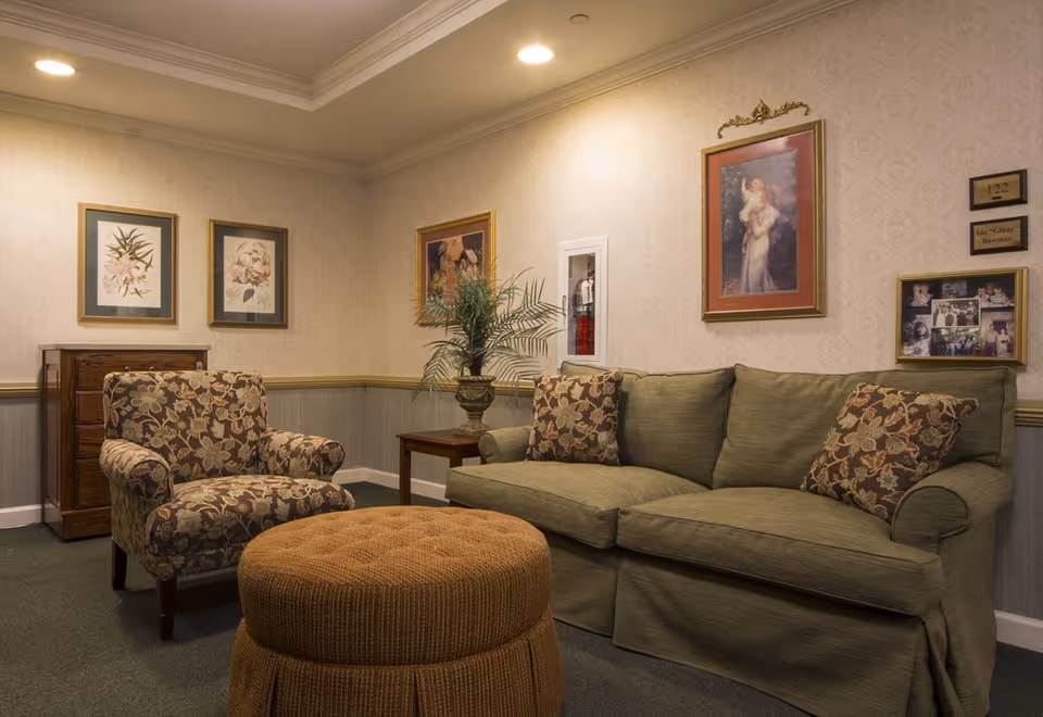 Cozy sitting area with a green sofa, patterned armchair, round ottoman, side table and framed artwork on the walls.