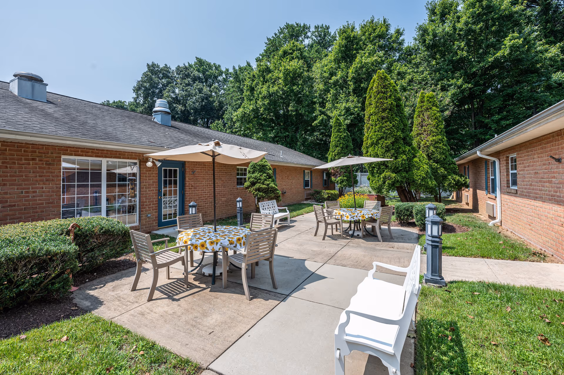 Outdoor patio area at Arden Courts - ProMedica Memory Care Community (Potomac) featuring two tables with sunflower-patterned tablecloths, each surrounded by chairs and shaded by umbrellas. The patio is bordered by brick buildings, green bushes, tall trees, and a white bench on the concrete walkway.