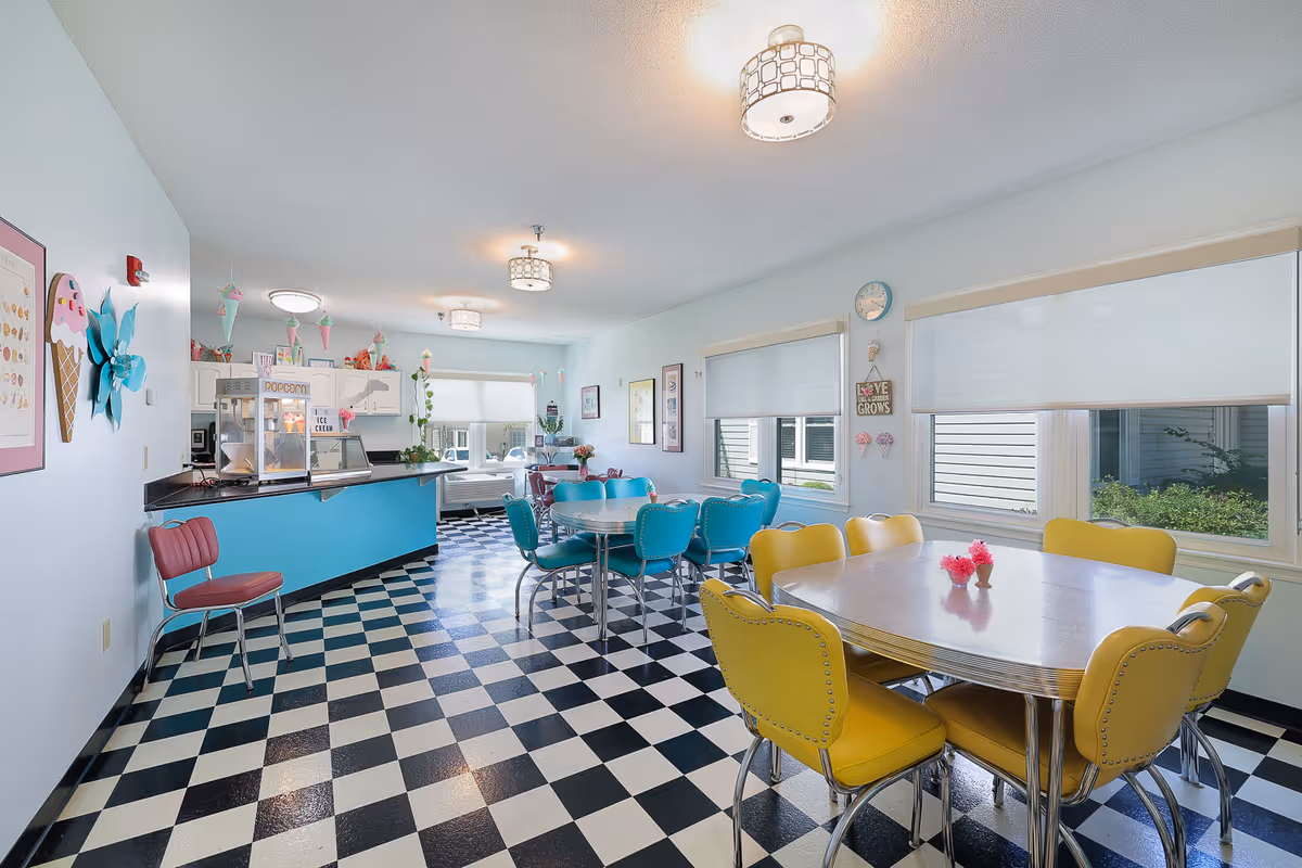 Bright and colorful dining room with retro-style furniture including yellow and turquoise chairs around tables. The floor has a black and white checkered pattern. The walls are decorated with ice cream and popcorn themed decorations. Large windows with white roller shades let in natural light. A popcorn machine is visible on the counter in the background.
