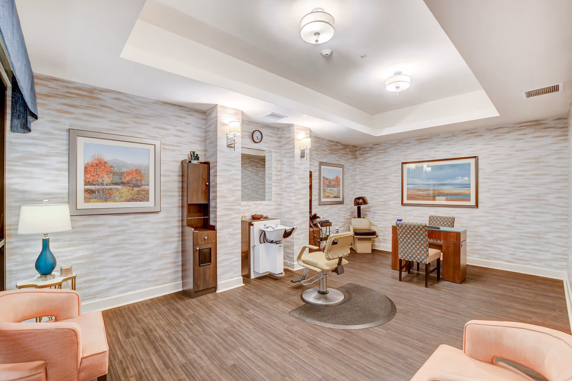 Well-lit interior salon-style common room with a barber chair, manicure table, seating, and framed artwork on the walls.