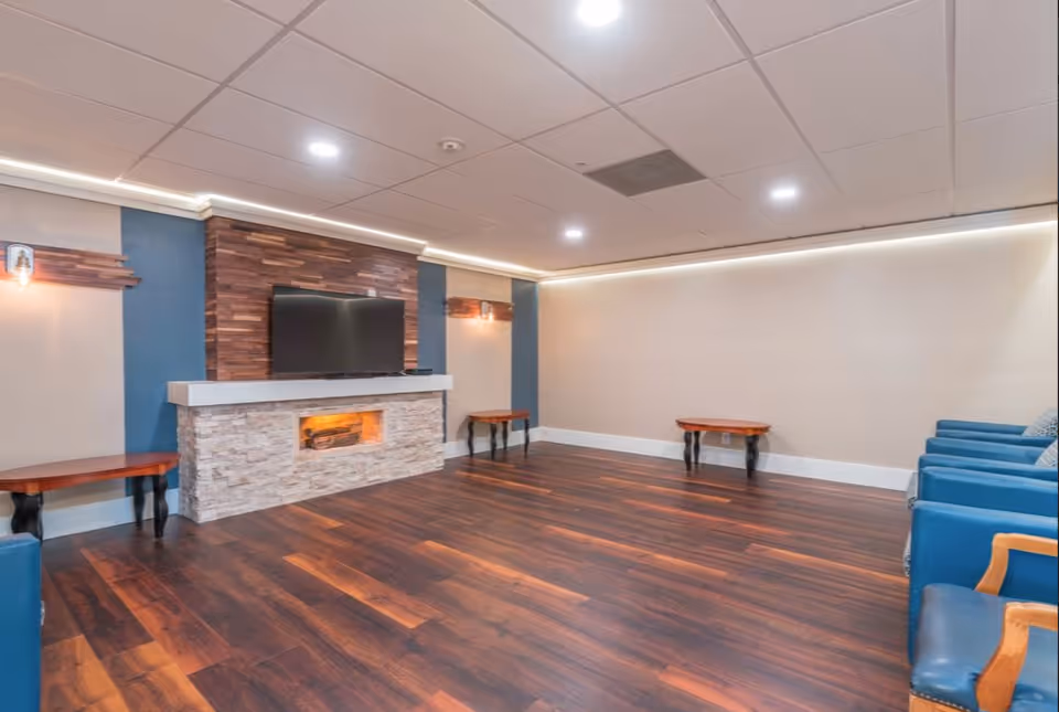 A spacious living room with dark wood flooring, a modern fireplace with a stone facade, a mounted flat-screen TV above the fireplace, blue armchairs lined up along the right wall, and small wooden side tables against the walls. The ceiling has recessed lighting and a white drop ceiling with LED strip lighting along the perimeter.