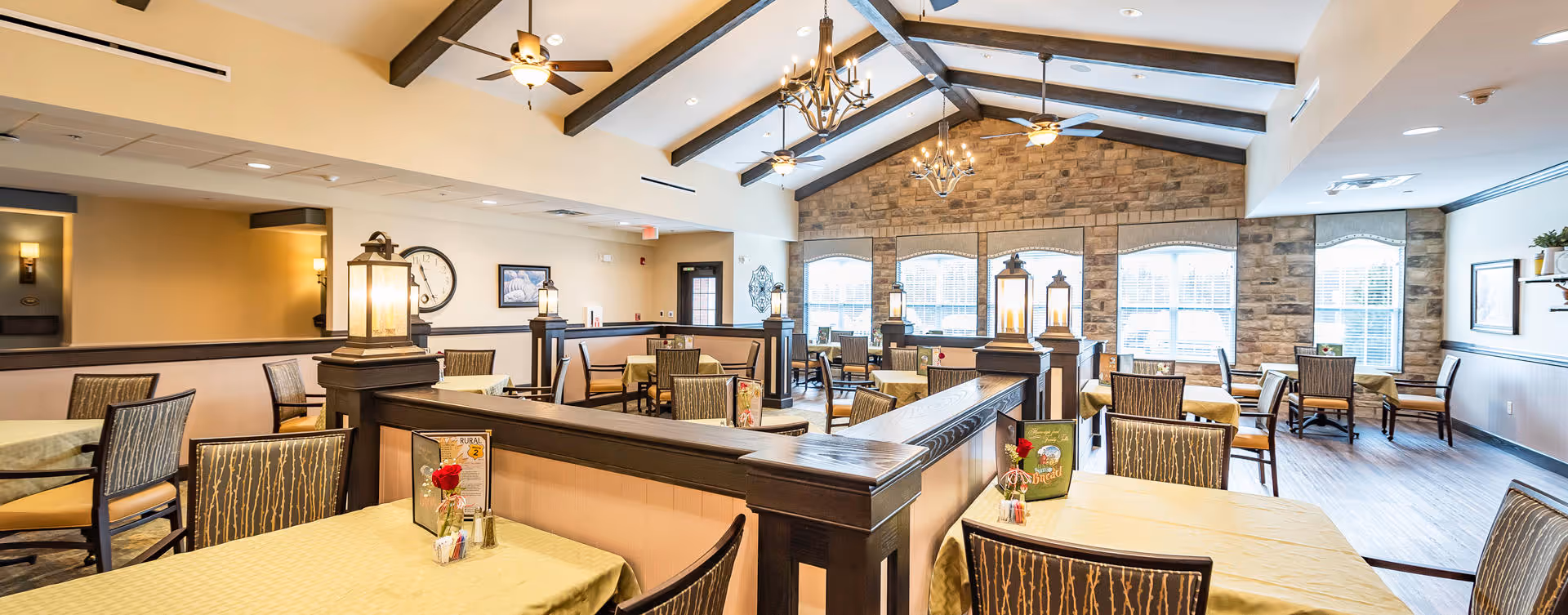 A spacious dining room with multiple tables covered in yellow tablecloths, each set with chairs and small flower centerpieces. The room features high vaulted ceilings with exposed beams, ceiling fans, and large windows letting in natural light. The walls have a combination of stone and light-colored paint, and there are decorative lantern-style lights on wooden dividers between seating areas.