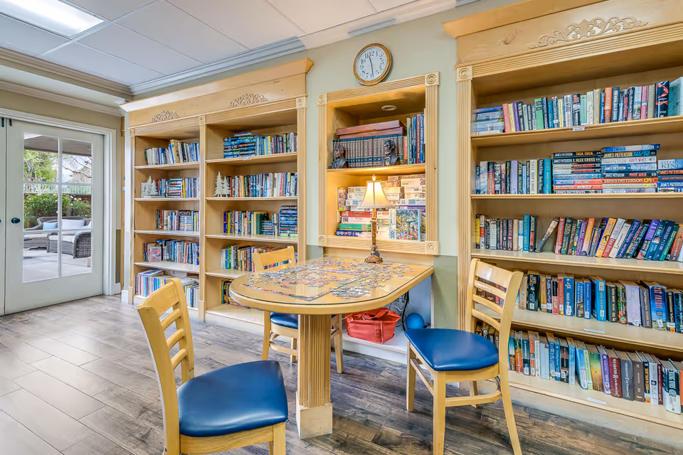 A cozy reading and puzzle area in a senior living facility with wooden bookshelves filled with books, a table with a partially completed jigsaw puzzle, and three wooden chairs with blue cushions. A small lamp is on the table, and a clock is mounted on the wall above the shelves. Glass doors lead to an outdoor patio with seating.