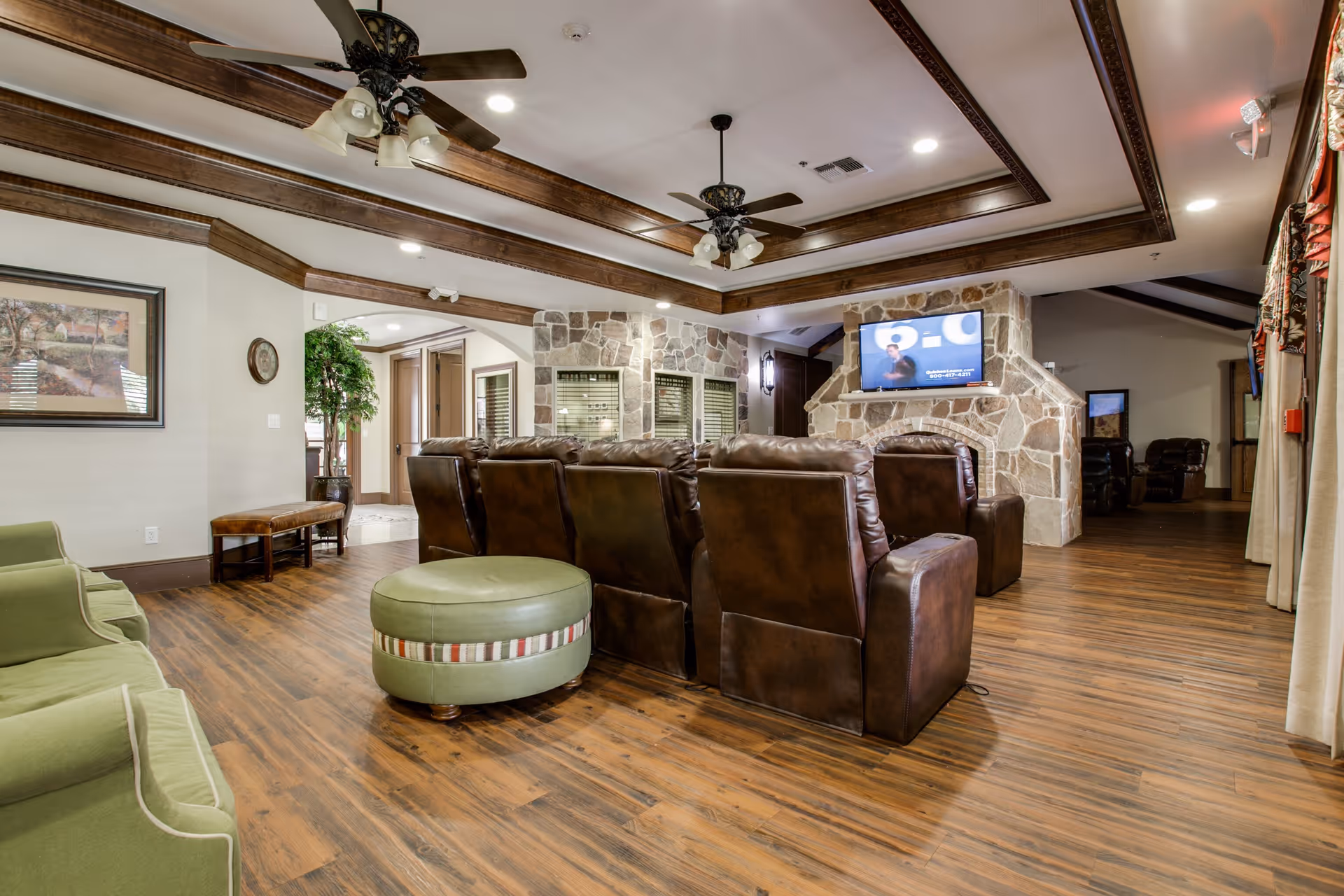 A cozy living room area in Avalon Memory Care featuring a row of brown leather recliners facing a wall-mounted TV above a stone fireplace. The room has wooden flooring, ceiling fans with lights, green upholstered chairs, a round green ottoman, and decorative wooden ceiling beams. There is a framed picture on the wall and a potted plant near an archway leading to another room.