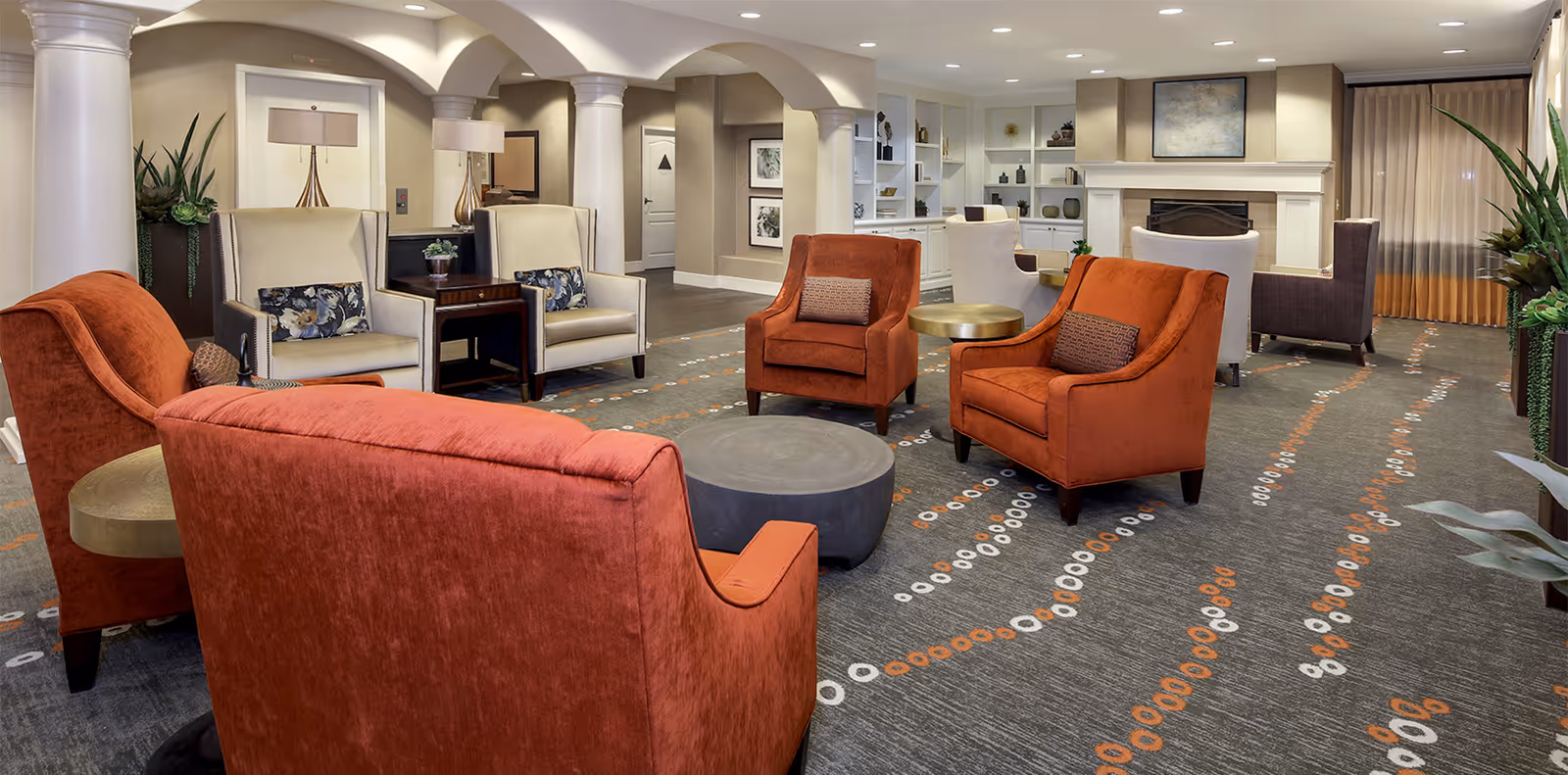A spacious and well-lit living room area with multiple comfortable armchairs in shades of orange and beige arranged around circular tables. The room features decorative columns, built-in shelving with various decor items, a fireplace, and large windows with curtains. The carpet has a modern pattern with orange and white circular designs, and there are several plants placed around the room.