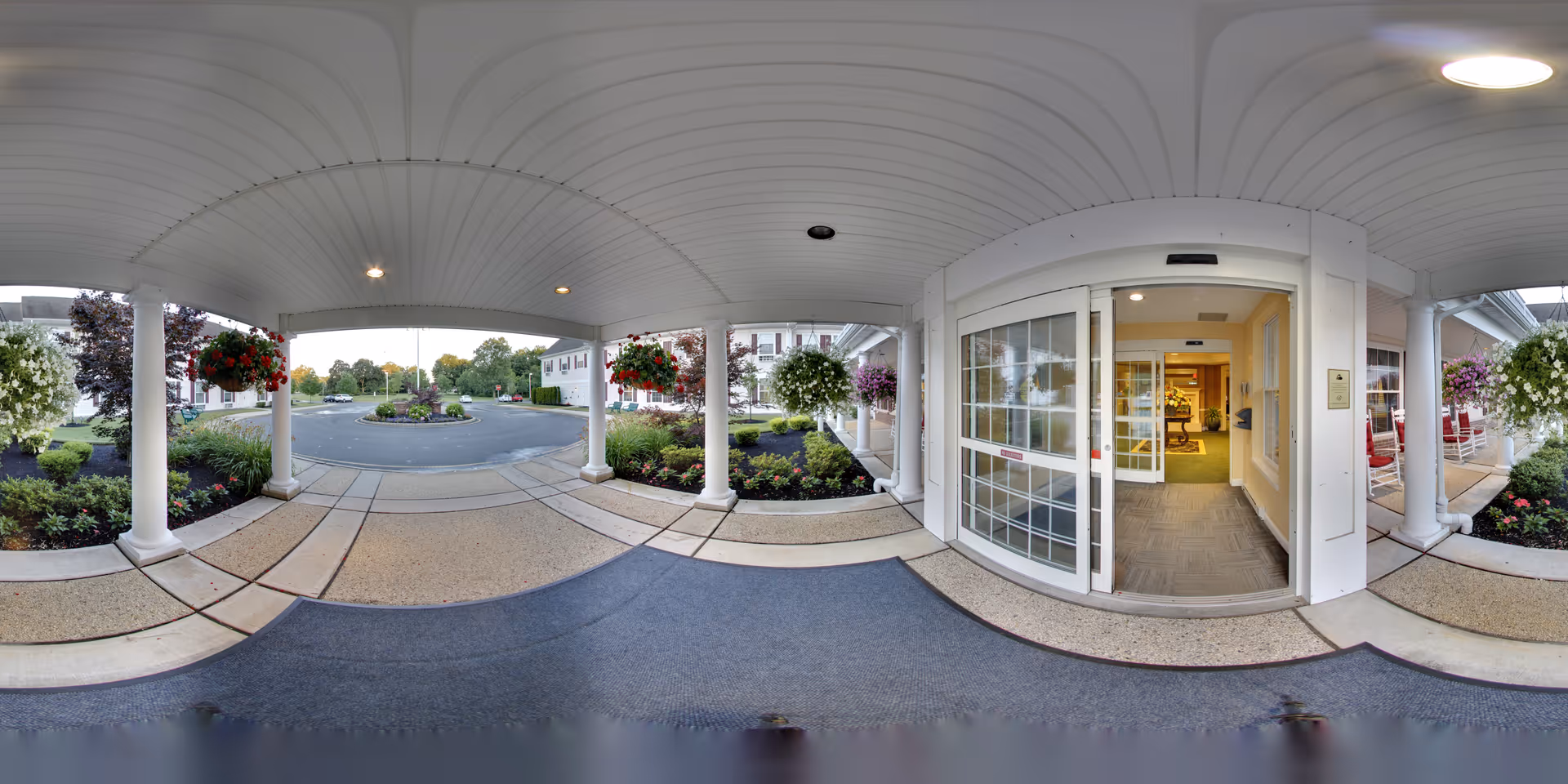 Covered entrance area of Brandywine Toms River by Monarch with white pillars, hanging flower baskets, and a sliding glass door leading into the building. Outside, there is a circular driveway with landscaped greenery and trees in the background.