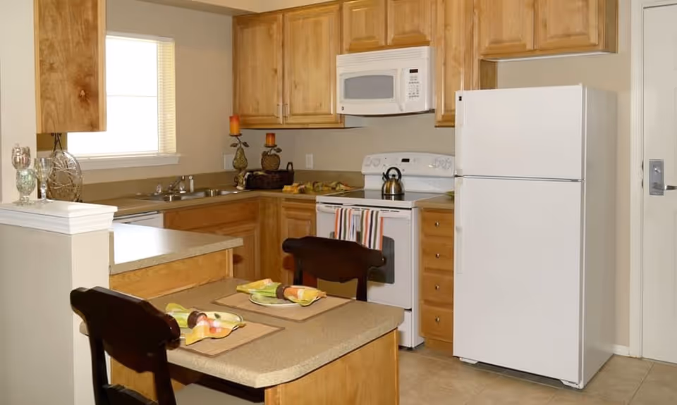 A kitchen area in a senior living facility featuring wooden cabinets, a white refrigerator, a white stove with a kettle on it, a white microwave above the stove, a double sink under a window, and a small dining counter with two place settings and dark wooden chairs.