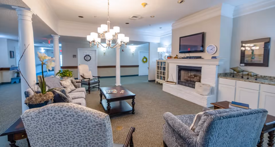 A spacious and well-lit living room area in a senior living facility featuring comfortable upholstered chairs and sofas arranged around a dark wooden coffee table. The room includes white pillars, a white brick fireplace with a mounted flat-screen TV above it, a clock, and decorative items on the mantel. There is a chandelier hanging from the ceiling, carpeted flooring, and built-in cabinets with a granite countertop on the right side. The walls are painted light beige, and there are doors and wall decorations visible in the background.