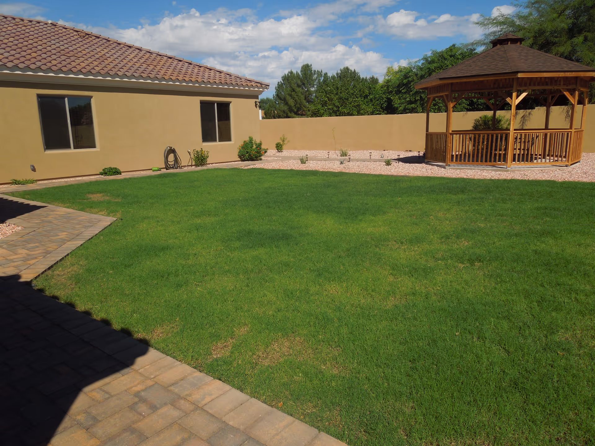 A well-maintained outdoor area of a facility featuring a green lawn, a paved walkway, a beige building with two windows, and a wooden gazebo under a partly cloudy sky.