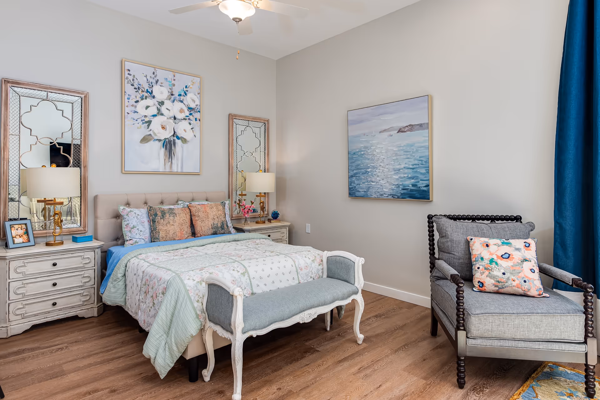 A cozy bedroom with a bed featuring floral and patterned bedding, two bedside tables with lamps and decorative mirrors above them, a cushioned bench at the foot of the bed, a gray armchair with a colorful floral pillow, and two paintings on the walls. The room has wooden flooring and a ceiling fan.
