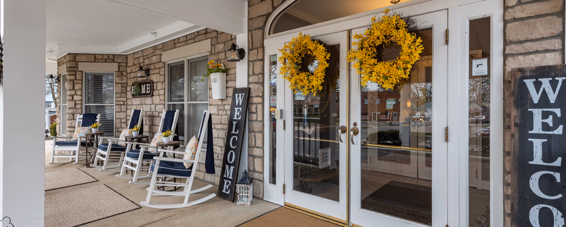 Covered front porch with rocking chairs, welcome signs, and double glass doors adorned with yellow wreaths.