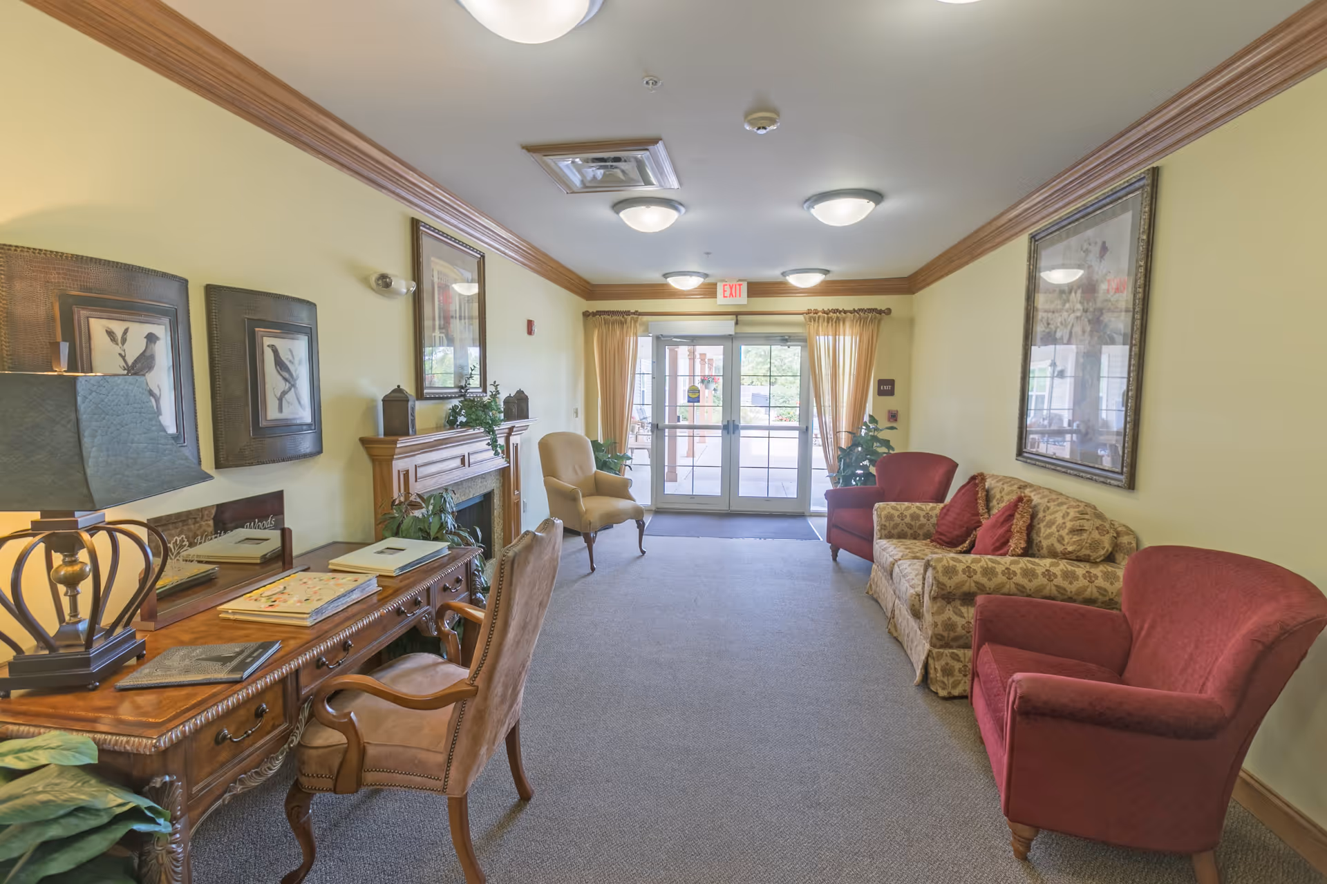 A cozy interior common area with a wooden desk and chair on the left, a fireplace with decorative items above it, and several upholstered chairs and a patterned sofa on the right. The room has light yellow walls with wooden crown molding, framed artwork, and a glass double door at the far end with curtains, leading outside.
