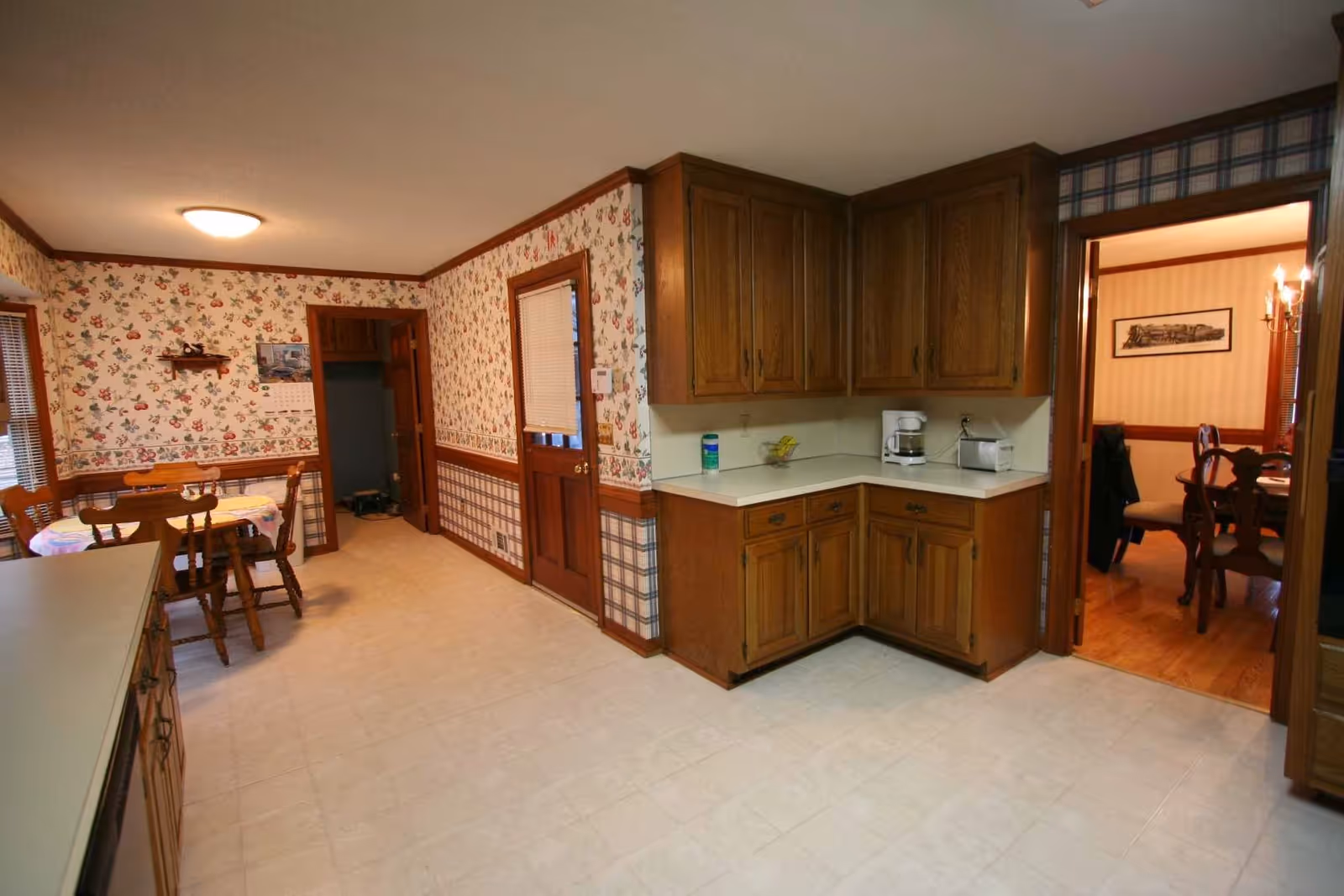 Interior view of a kitchen with wooden cabinets, white countertops, and a small dining table with chairs. The walls are decorated with floral and plaid wallpaper. There is a door leading to another room with a wooden dining table and chairs visible.