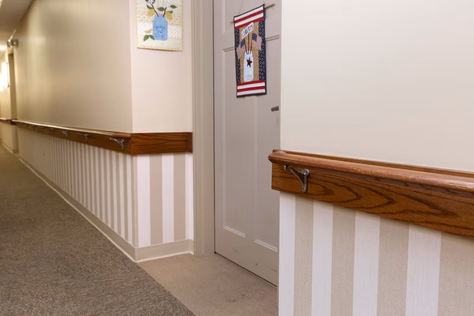 A hallway in a senior living facility with beige and white striped wallpaper on the lower half of the walls, wooden handrails, and a closed door decorated with a patriotic hanging that says 'Liberty'. The hallway is carpeted and well-lit with wall-mounted lights.