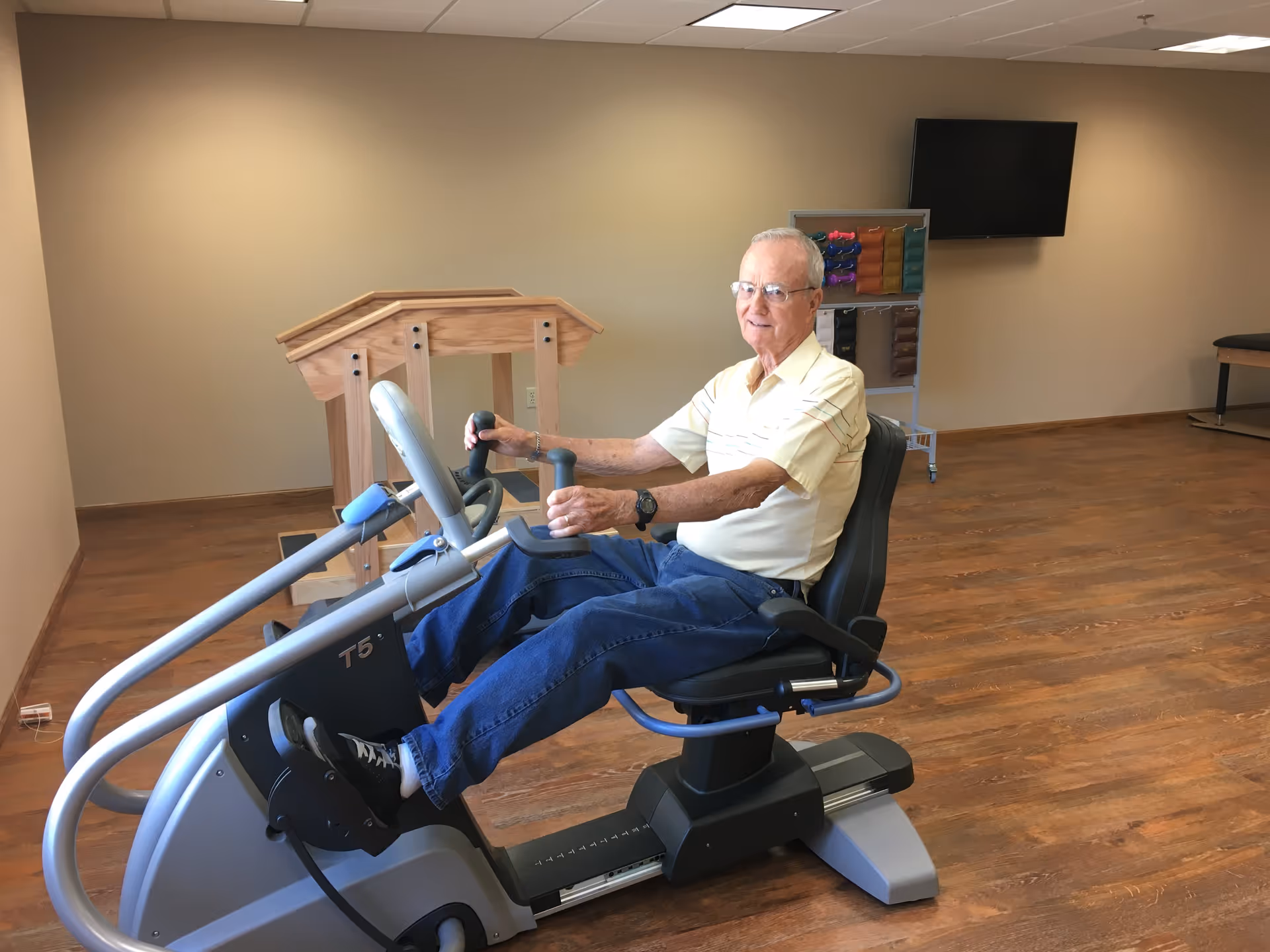An elderly man wearing glasses, a light yellow shirt, and blue jeans is seated on a recumbent exercise bike in a spacious room with wooden flooring. Behind him, there is a wooden handrail structure, a rack with colorful exercise bands, and a wall-mounted flat-screen TV.