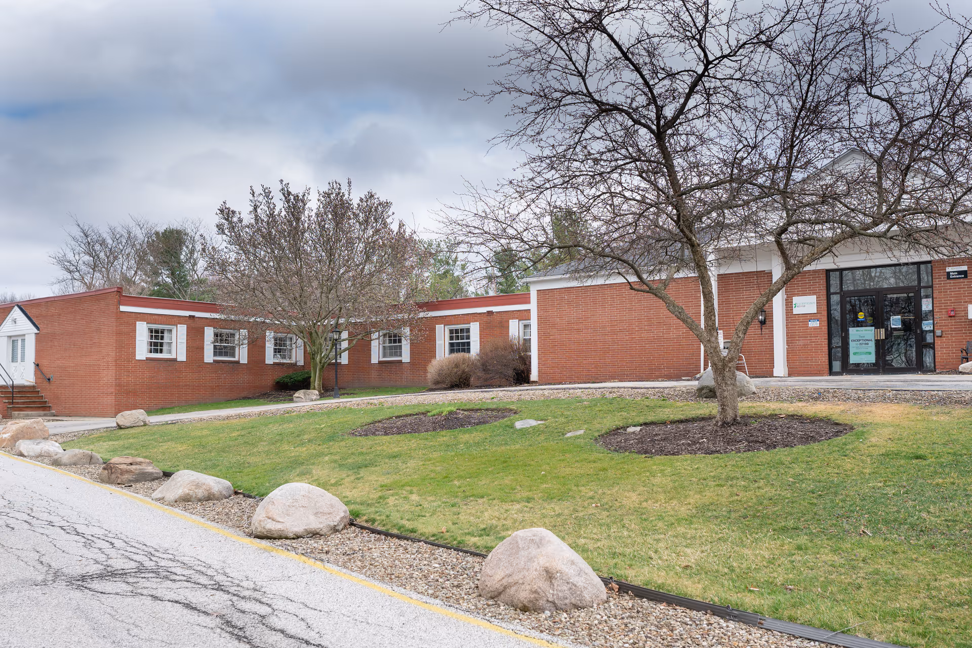 Front exterior of a low red-brick building with a lawn, leafless trees, and a glass main entrance.