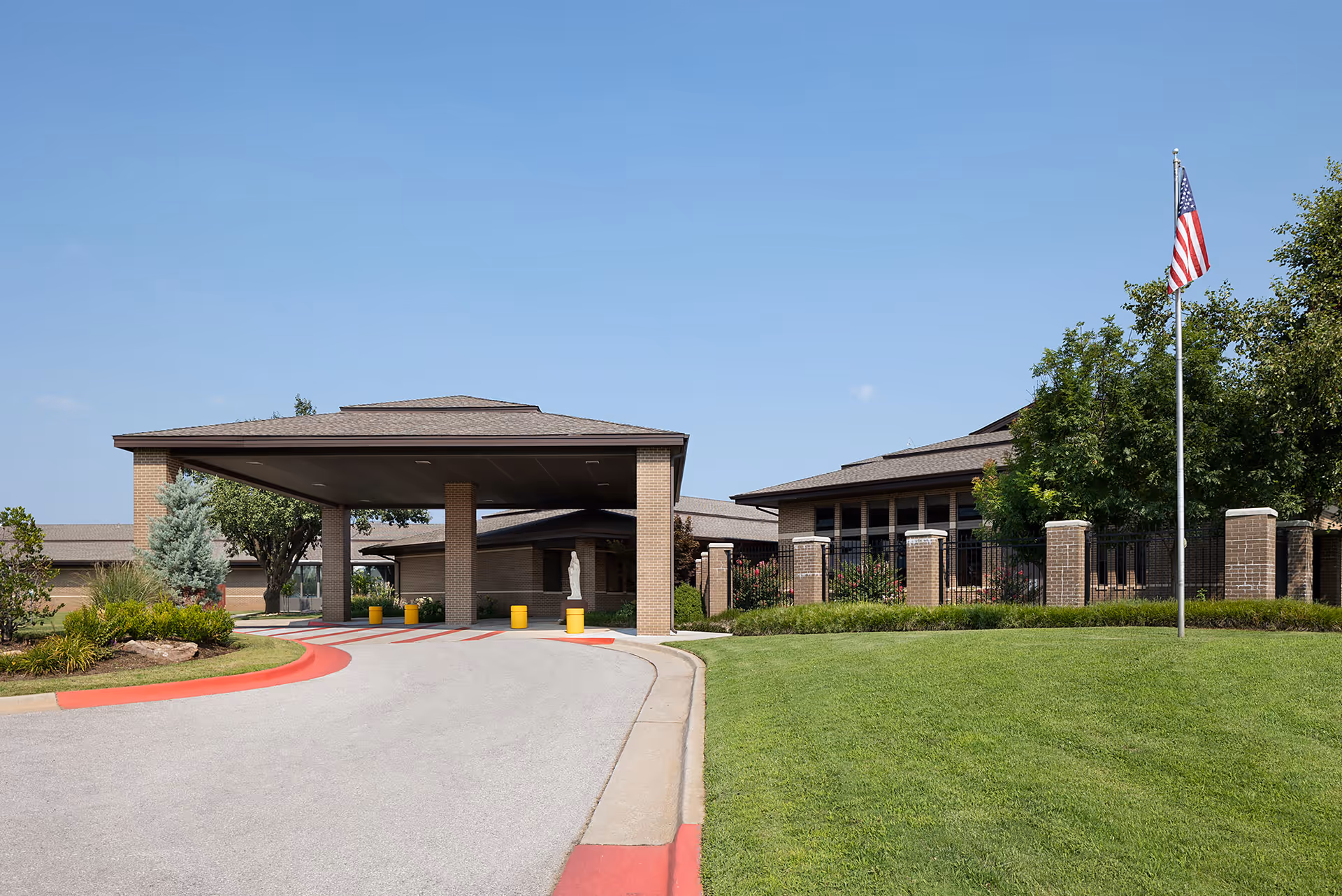 Exterior view of St. Ann's Skilled Nursing & Therapy facility showing the entrance with a covered drop-off area, a driveway with red curbs, green lawn, trees, and an American flag on a flagpole under a clear blue sky.