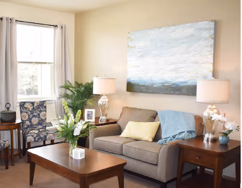 A cozy living room with a beige sofa adorned with a yellow pillow and a blue throw blanket. Two wooden side tables with matching lamps flank the sofa. A wooden coffee table with a floral arrangement sits in front of the sofa. A patterned armchair is positioned near a window with light curtains. A large abstract painting hangs on the wall above the sofa.